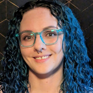 A white woman in her early 30s has dark, curly blue hair, a septum piercing, and square aqua glasses.