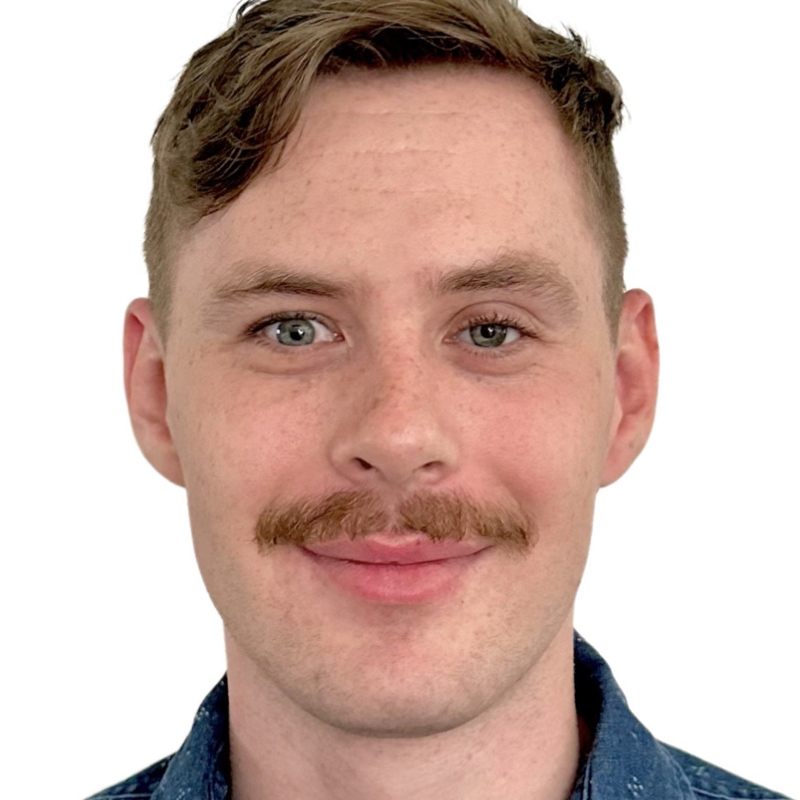 A white man in his late 20s wears a denim collared shirt. His dark blond hair is styled in a "more on top" haircut, and his trimmed mustache lines his upper lip as he smiles.
